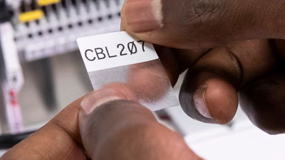 Tecnico che sta per applicare un’etichetta in vinile autolaminante Brady con adesivo aggressivo su un filo o un cavo. Il testo è stampato in modo chiaro sulla sezione bianca dell’etichetta, mentre una sezione trasparente avvolge e copre il testo una volta applicata.