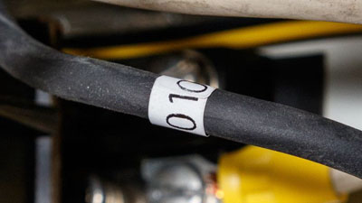 Etichetta wrap around in tedlar bianco autoestinguente con testo nero, applicata intorno a un filo nero di un quadro elettrico.